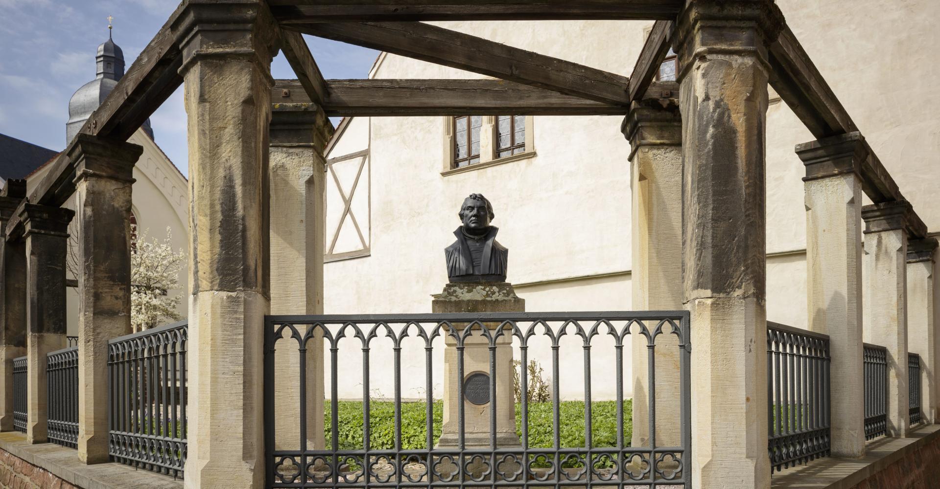 Büste Luthers im Fokus, Zaun davor, im Hintergrund Hauswand