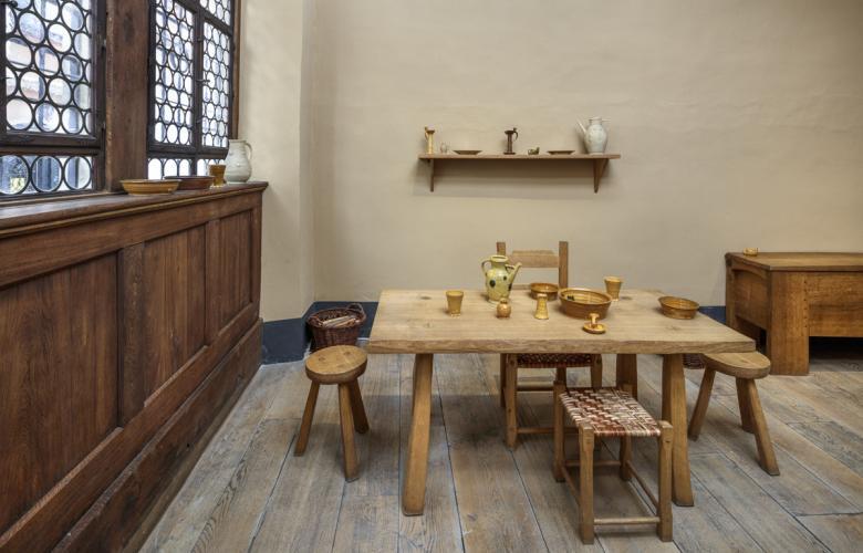 nachgestellte Stube. Holztisch mit 4 Hockern, helle Wand, dunkle Holzfenster und heller Fußboden